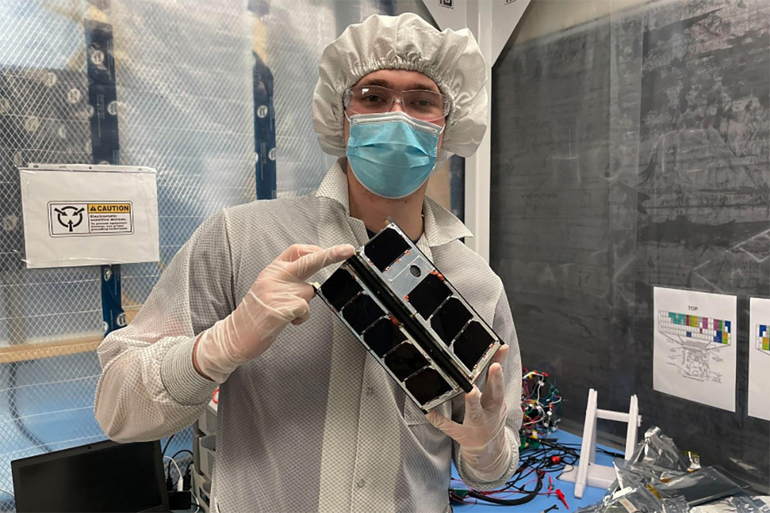 A person in white hazmat clothing holds up a rectangular shaped satellite about the size of a kleenex box. 