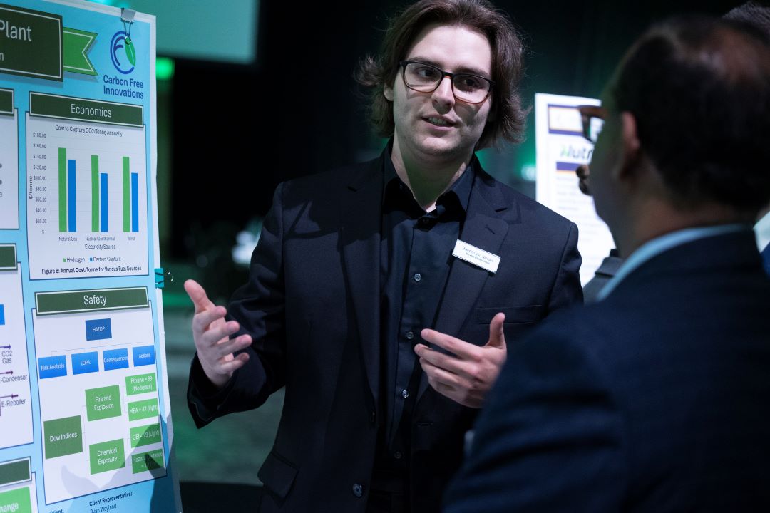 Male engineering student standing in front of a poster is speaking to a middle-aged man. 